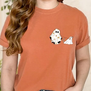 May include: A burnt orange t-shirt featuring a white ghost wearing a pattern of orange flowers and holding a leash attached to a smaller white ghost dog.
