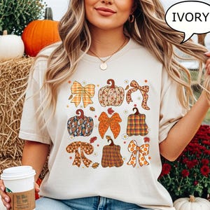 Könnte beinhalten: Cremefarbenes T-Shirt mit einem Design aus illustrierten Kürbissen und Schleifen in verschiedenen Mustern und Farben. Das Design umfasst Orange-, Braun- und Gelbtöne mit floralen und karierten Details. Das Shirt ist ein lässiges Kleidungsstück zum Thema Herbst.