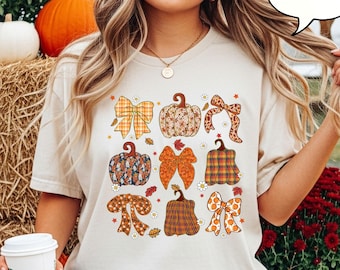 May include: Ivory-colored t-shirt featuring a collection of illustrated pumpkins and bows in various patterns and colors. The design includes orange, brown, and yellow hues, with floral and plaid details. The shirt is a casual, fall-themed apparel item.