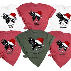 Peut inclure: Six t-shirts rouges, blancs et verts avec une silhouette noire et blanche de Mickey Mouse portant un bonnet de P&egrave;re No&euml;l. La silhouette est devant une image noire et blanche du ch&acirc;teau de Cendrillon avec des feux d'artifice. Le texte "2025" est au-dessus de la silhouette. Chaque t-shirt a un mot diff&eacute;rent sous la silhouette : "Papa", "Maman", "Fr&egrave;re", "S&oelig;ur", "Nom" et "Tante".