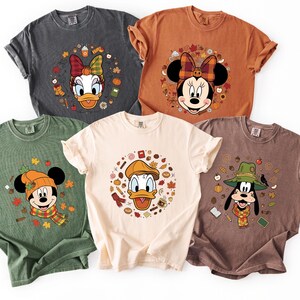 May include: Five short-sleeved t-shirts in various colors, each featuring a different Disney character in a fall-themed design. Characters include Daisy Duck, Minnie Mouse, Mickey Mouse, Donald Duck, and Goofy, surrounded by autumn leaves and accessories.