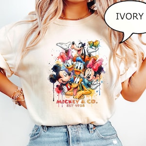 May include: Ivory-colored t-shirt featuring a colorful watercolor design of Mickey Mouse, Minnie Mouse, Donald Duck, Daisy Duck, Goofy, and Pluto. The shirt includes the text "Mickey & Co. EST 1928".