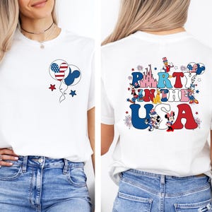 K&ouml;nnte beinhalten: Wei&szlig;es T-Shirt mit patriotischem Design. Die Vorderseite zeigt Mickey Mouse-f&ouml;rmige Ballons in Rot, Wei&szlig; und Blau, mit Sternen. Auf der R&uuml;ckseite steht der Text "Party in the USA" mit Mickey- und Minnie-Mouse-Figuren.