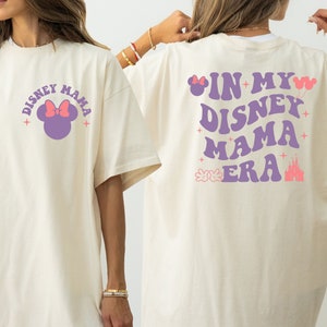 May include: A cream-coloured t-shirt featuring the text "DISNEY MAMA" and a Minnie Mouse graphic on the front. The back of the shirt displays the phrase "IN MY DISNEY MAMA ERA" in purple, accompanied by Disney-themed graphics, including a castle and balloons.