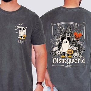 Pu&ograve; includere: T-shirt grigio scuro con un design a tema Halloween. La parte anteriore mostra un personaggio fantasma con una zucca e la parola "NAME". Il retro presenta una scena di casa infestata con la scritta "Disneyworld est.1971".