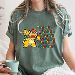 May include: Green t-shirt featuring a cartoon Bowser character holding a microphone. The text "PEACHES I LOVE YOU" is printed in colorful letters and musical notes. The shirt is a comfortable, casual style.