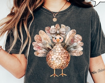 Könnte beinhalten: Dunkelgraues T-Shirt mit einem farbenfrohen Truthahn-Motiv, einer karierten Schleife, Glitzer und Federdetails. Das Wort "PEPPER" steht in einer Sprechblase. Das Shirt ist ein bequemer, lässiger Stil.