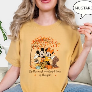May include: Mustard-colored t-shirt featuring Mickey and Minnie Mouse under a tree with autumn leaves. The graphic includes pumpkins, a boot, and the text "It's the most wonderful time of the year."