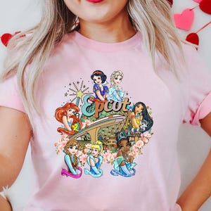 May include: Pink t-shirt featuring a graphic of Disney princesses in front of the Epcot logo and Spaceship Earth. The princesses include Snow White, Ariel, Belle, Cinderella, Pocahontas, Elsa, and Anna.