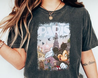 May include: A dark gray t-shirt with a vintage-style graphic of Mickey Mouse in front of the Eiffel Tower in Paris. The graphic includes the text "Paris" and "Bon Voyage!"  The shirt has a distressed look and a crew neck.