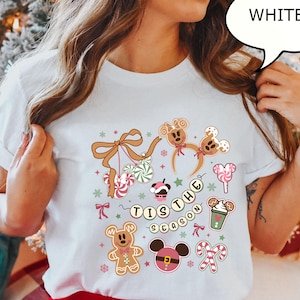 May include: White t-shirt with a festive design featuring gingerbread men, candy canes, and the phrase "Tis the Season." The design includes a Mickey Mouse-inspired gingerbread cookie and a cup of hot cocoa.