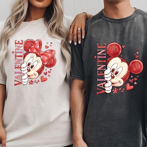 Puede incluir: Dos camisetas holgadas, una crema y otra carb&oacute;n, con un dise&ntilde;o de San Valent&iacute;n. El dise&ntilde;o incluye un rat&oacute;n de dibujos animados con un lazo rojo y corazones, con la palabra "VALENTINE" impresa verticalmente en rojo.