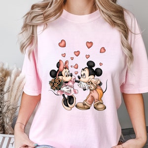 May include: Pink t-shirt with a graphic of Mickey Mouse and Minnie Mouse holding hands with hearts floating around them.