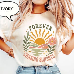 May include: An ivory t-shirt featuring a graphic print of a sun rising over hills and water, flanked by a leafy branch and a flower. The text "FOREVER" is arched above the sun, and "CHASING SUNSETS" is printed in a curved line below the graphic.