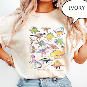 May include: Ivory-colored t-shirt featuring a colorful dinosaur graphic. The graphic includes various dinosaur illustrations with their names, such as Tyrannosaurus and Stegosaurus. The shirt is a casual style, perfect for a dinosaur enthusiast.