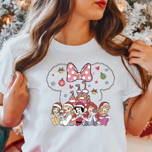 Puede incluir: Camiseta blanca con un dise&ntilde;o navide&ntilde;o con un lazo rosa, un castillo y seis princesas Disney con gorros de Pap&aacute; Noel. Las princesas sostienen objetos navide&ntilde;os.