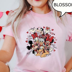 May include: Pale pink t-shirt with a Disney character graphic including Mickey and Minnie Mouse, Donald and Daisy Duck, Goofy, and Pluto, with hearts. The shirt has rolled sleeves and a speech bubble with the word "BLOSSOM".