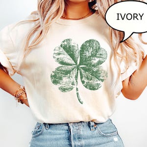 Könnte beinhalten: Elfenbeinfarbenes T-Shirt mit einem verblassten grünen vierblättrigen Kleeblatt-Motiv. Das Shirt hat eine lockere Passform und hochgekrempelte Ärmel. Das Wort "IVORY" steht in einer Sprechblase. Das Shirt wird mit blauen Jeans kombiniert.