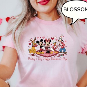 Puede incluir: Camiseta rosa claro con un dise&ntilde;o de San Valent&iacute;n que presenta a los personajes de Disney Mickey, Minnie, Donald, Goofy y Pluto. La frase "Mickey's Very Happy Valentine's Day" est&aacute; impresa debajo de los personajes. Tambi&eacute;n es visible un bocadillo con la palabra "BLOSSOM".