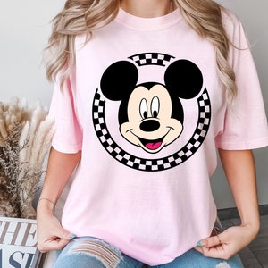 Puede incluir: Una camiseta rosa con un c&iacute;rculo de cuadros negros y blancos alrededor de una imagen de dibujos animados de Mickey Mouse. Mickey Mouse est&aacute; sonriendo con la lengua fuera.