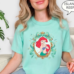 May include: A light teal t-shirt featuring a framed illustration of a red-haired mermaid with a blue seashell top and green tail. The frame is gold with teal ribbons and floral accents. The text "ISLAND REEF" is in a speech bubble.