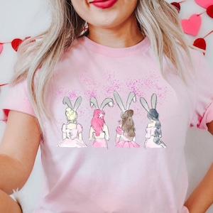 Puede incluir: Camiseta rosa con un gr&aacute;fico de cuatro mujeres con orejas de conejo y vestidos rosas. El gr&aacute;fico est&aacute; rodeado de purpurina rosa.
