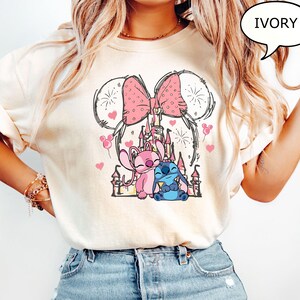 Puede incluir: Camiseta de color marfil con un gr&aacute;fico de dibujos animados de un lazo rosa con lunares, orejas de Mickey Mouse y un castillo. El gr&aacute;fico incluye corazones rosas, fuegos artificiales y dos personajes de dibujos animados. La palabra "IVORY" est&aacute; en una burbuja.