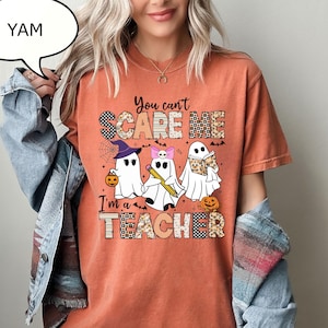 May include: A burnt orange short-sleeve t-shirt featuring a graphic of three ghosts with the text "You can't SCARE ME I'm a TEACHER". The ghosts are depicted with a witch hat, a pink bow, and a book, alongside small pumpkins and bats.