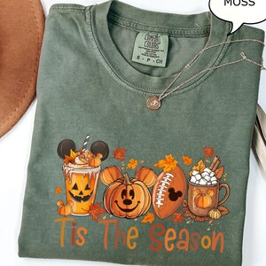 May include: A moss green Comfort Colors t-shirt with a fall-themed graphic. The graphic features a pumpkin spice latte with Mickey Mouse ears, a pumpkin with a Mickey Mouse face, a football with a Mickey Mouse head, and a mug of marshmallows. The words 'Tis The Season' are printed in orange and brown.