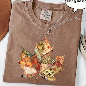 May include: Espresso-colored Comfort Colors t-shirt with a large, patchwork leaf design. The leaf features various fabric patterns in fall colors. A silver necklace with a small pendant is draped over the shirt. The shirt is size S-P-CH.