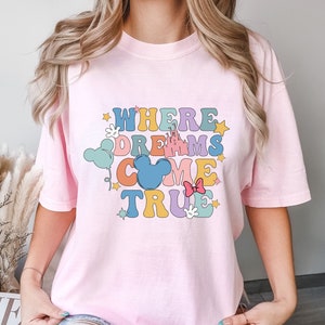 May include: Pink t-shirt with a graphic design that says "Where Dreams Come True" in colorful letters with a Mickey Mouse head and other Disney-themed icons.
