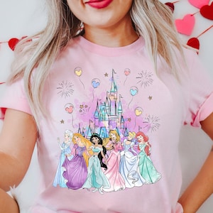 Puede incluir: Camiseta rosa con un gr&aacute;fico de princesas Disney de pie frente al castillo de Cenicienta con fuegos artificiales y globos.