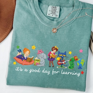 Puede incluir: Una camiseta de manga corta en color verde salvia con un gráfico de varios personajes de libros infantiles leyendo y tocando instrumentos. Debajo de los personajes, el texto blanco dice, it's a good day for learning. Etiqueta Comfort Colors.