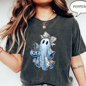 May include: Dark grey t-shirt with a ghost design. The ghost is light blue with patches and a floral bow, holding a blue pumpkin bucket. The word "PEPPER" is in a speech bubble.