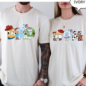 May include: Ivory t-shirts with the words "DAD" and "MOM" in colourful, cartoon-style lettering. The "DAD" shirt features characters and imagery from a popular animated film. The "MOM" shirt has similar design elements.