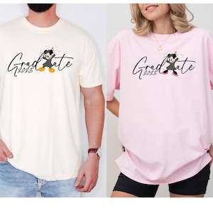 Puede incluir: Dos camisetas con un tema de graduaci&oacute;n. Una es de color crema y la otra es rosa. Ambas camisetas presentan una imagen de dibujos animados de Mickey Mouse con una gorra y una bata de graduaci&oacute;n. El texto "Graduate 2023" est&aacute; impreso en ambas camisetas.