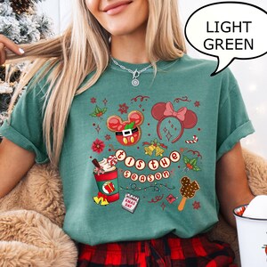 May include: Light green t-shirt with a festive graphic featuring Mickey Mouse-themed Christmas elements. The design includes a gingerbread cookie, a bow, a coffee cup, and the text "'Tis the Season". The shirt is paired with red and black plaid pajama bottoms.