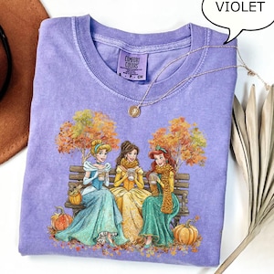 May include: Violet Comfort Colors t-shirt featuring a graphic of three princesses sitting on a bench, holding cups, with autumn trees and pumpkins. A gold necklace is draped over the shirt. The shirt is a light purple color.