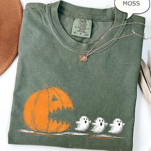 May include: A moss green t-shirt featuring a large orange jack-o'-lantern graphic with a carved mouth and three white ghost figures. The shirt has a Comfort Colors tag and a silver necklace with a pendant.