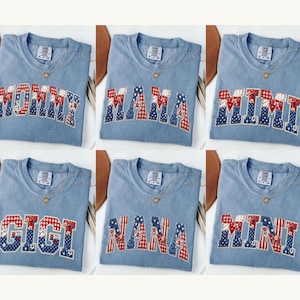 Puede incluir: Seis camisetas de color azul claro con las palabras "MOMMY", "MAMA", "MIMI", "GIGI", "NANA" y "MINI" en un diseño de patchwork con patrones rojos, blancos y azules. Cada camiseta tiene un collar dorado. Las camisetas se muestran sobre un fondo neutro.