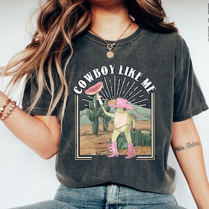 May include: A dark gray t-shirt with a vintage-style graphic of a frog wearing a cowboy hat and holding a lasso. The text "COWBOY LIKE ME" is printed above the frog.