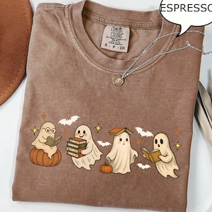 Könnte beinhalten: Espressofarbenes T-Shirt mit einer Grafik von Cartoon-Geistern, die Bücher lesen, auf Kürbissen sitzen und von Fledermäusen und Sternen umgeben sind. Das Shirt hat einen Rundhalsausschnitt und kurze Ärmel. Eine silberne Halskette liegt auf dem Shirt.