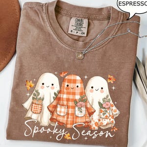 May include: Espresso-coloured t-shirt featuring a graphic of three friendly ghosts in floral and plaid outfits. The words "Spooky Season" are printed below. A silver necklace with a pendant is draped across the shirt.
