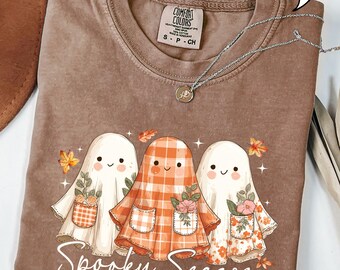 May include: Espresso-colored t-shirt with a graphic of three friendly ghosts wearing floral and plaid outfits. The phrase "Spooky Season" is printed below the ghosts. A silver necklace with a pendant is draped across the shirt.