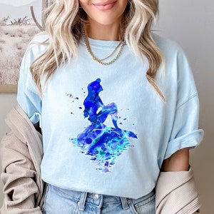 Puede incluir: Una camiseta azul claro con un dise&ntilde;o de acuarela de Ariel, la Sirenita, sentada en una roca con su cola en el agua. El dise&ntilde;o est&aacute; en tonos azules y verdes.
