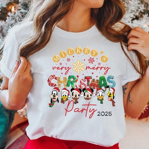 K&ouml;nnte beinhalten: Wei&szlig;es T-Shirt mit einem Mickey Mouse Weihnachtsparty-Design. Das Design zeigt Mickey Mouse, Minnie Mouse, Donald Duck, Goofy und Pluto, die Weihnachtsm&uuml;tzen und Weihnachtspullover tragen. Der Text auf dem Shirt lautet "Mickey's very merry Christmas Party 2025".