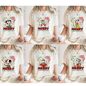 Puede incluir: Camisetas color crema con Mickey Mouse, el Pato Donald y Pluto en dise&ntilde;os navide&ntilde;os. Cada camiseta tiene una corona festiva en forma de coraz&oacute;n y las palabras "Merry Christmas" en rojo. Los personajes llevan gorros de Pap&aacute; Noel.