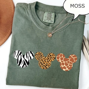 Pu&ograve; includere: T-shirt verde muschio con tre disegni a testa di Topolino con motivi zebrati, leopardati e giraffa. La maglietta ha girocollo e maniche corte. Una collana d'argento con un ciondolo &egrave; drappeggiata sulla maglietta.