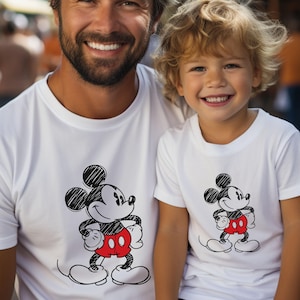 May include: A white t-shirt with a black and white sketch of Mickey Mouse wearing red shorts. The shirt is being worn by a man and a young boy.