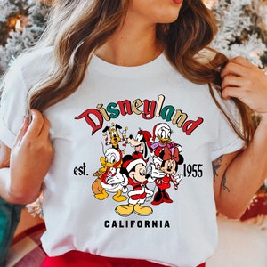 May include: White t-shirt with a colorful Disneyland logo featuring Mickey Mouse, Minnie Mouse, Donald Duck, Goofy, and Pluto wearing Santa hats. The text reads "Disneyland est. 1955 CALIFORNIA".